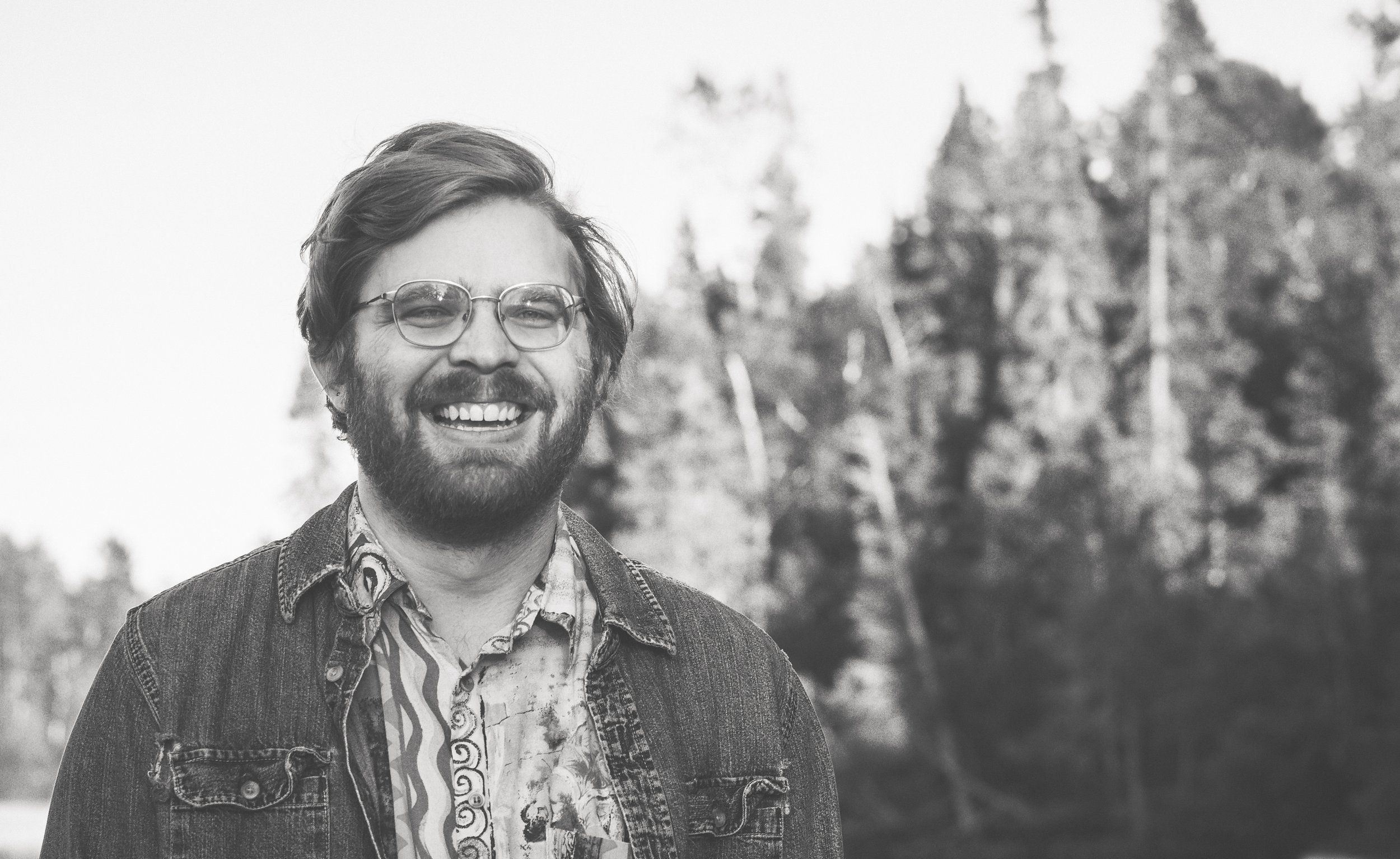 A black and white image of Ryan Ander-Evans smiling big with evergreen trees in the background. Ryan is white, wears glasses, has a beard, and has ear-length hair.