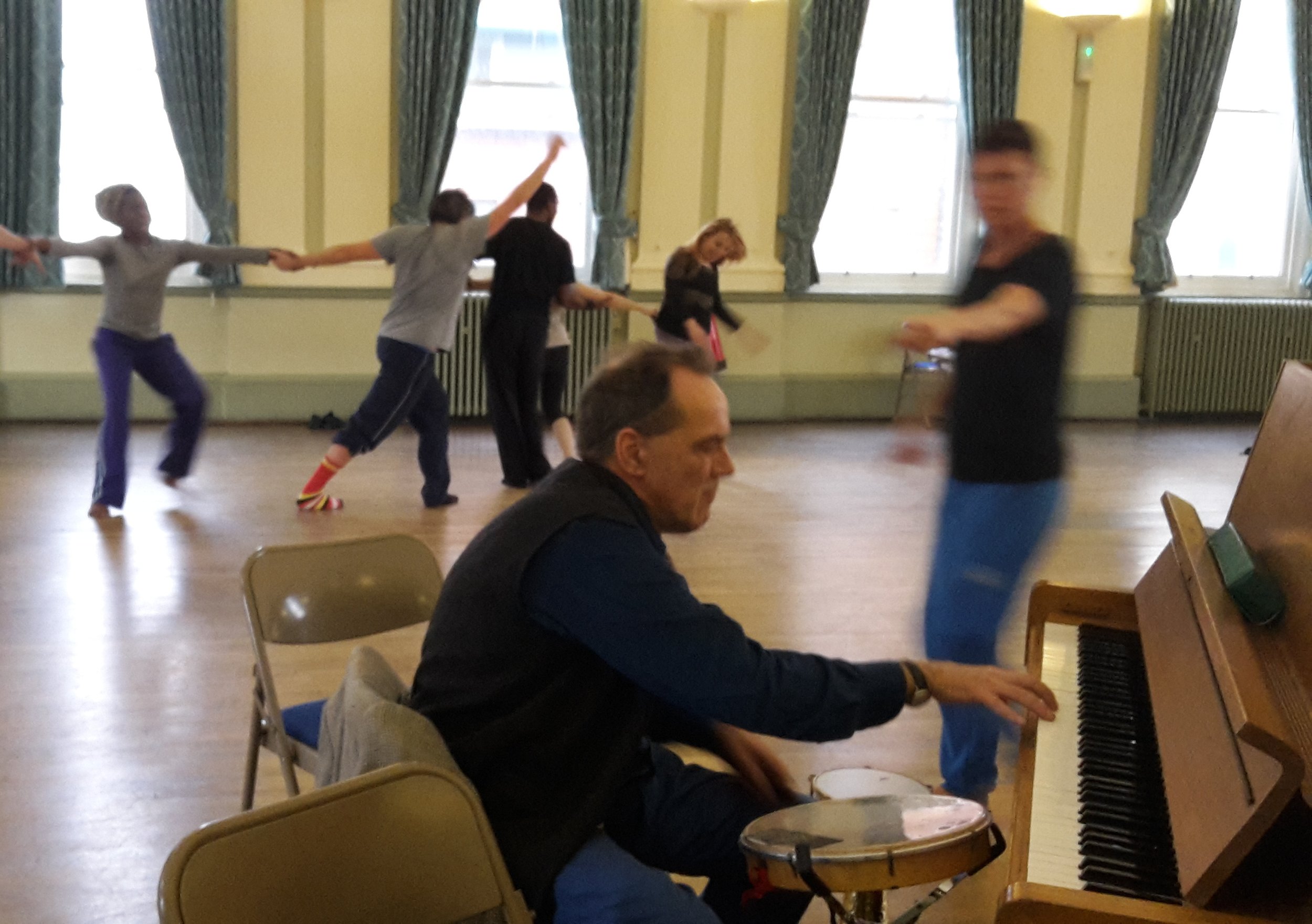 A seated person plays the piano while a group of people dance and move behind them in a well-lit room